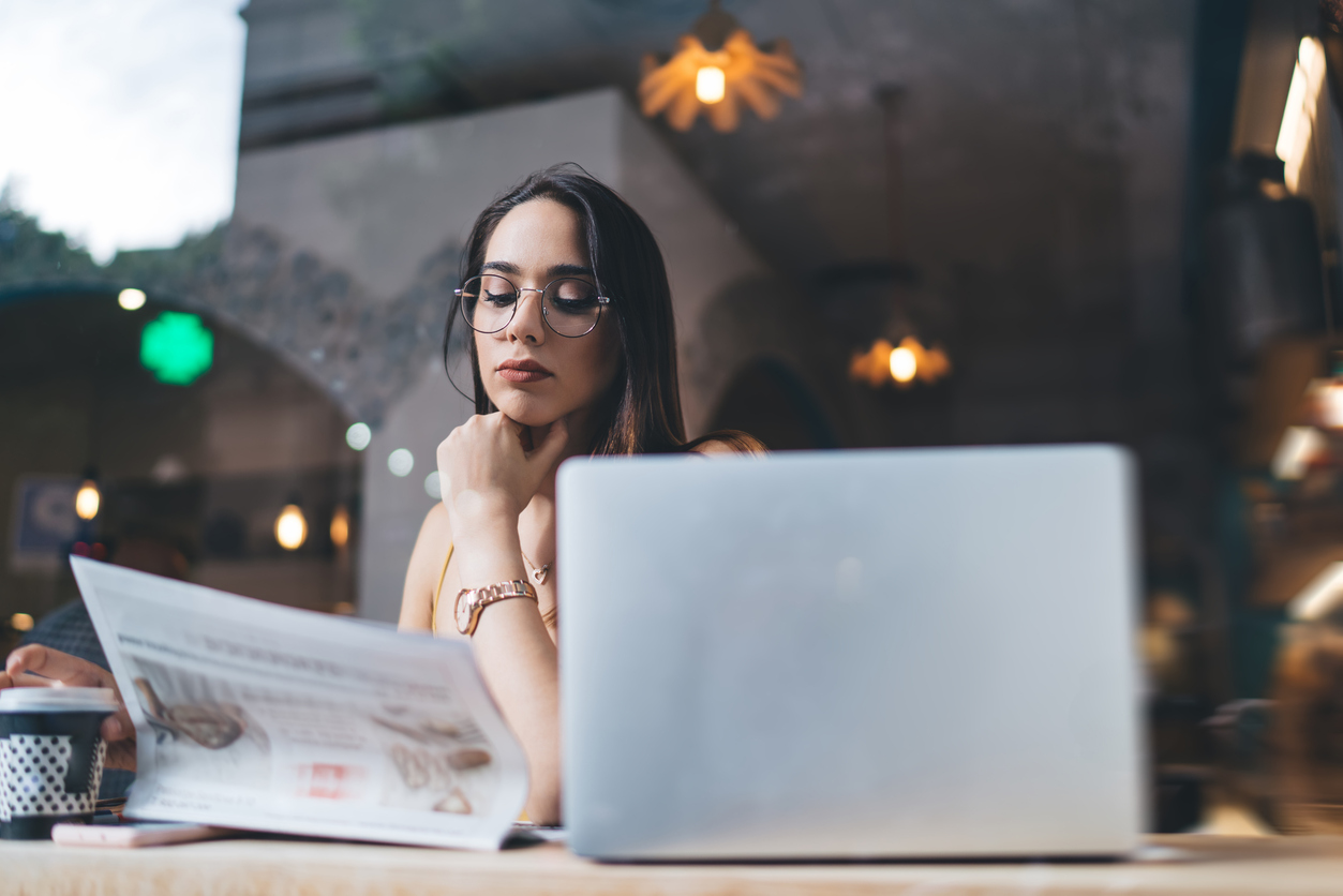 Through window of young serious woman in eyeglasses attentively reading newspaper and holding paper cup of coffee while sitting at table with laptop in cafe